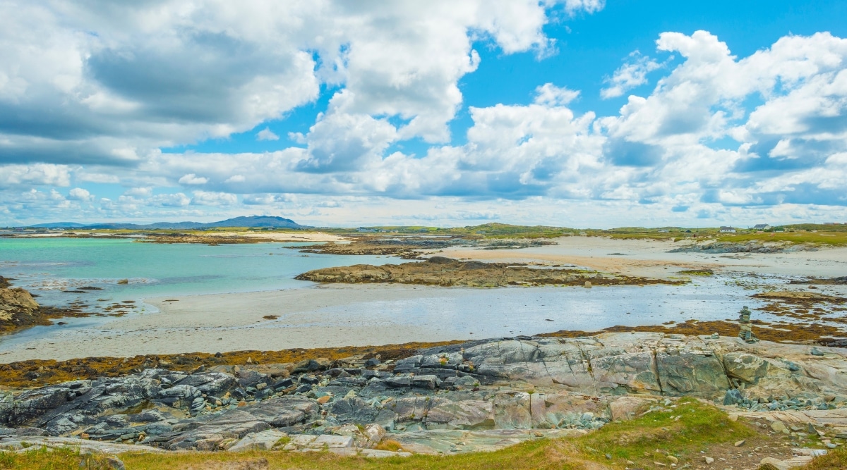 A photograph capturing Connemara Beaches in County Galway, Ireland. The image showcases the coastal landscape with sandy shores, and in the distance, the Atlantic Ocean meets the horizon. The beaches are characterized by natural elements, including scattered rocks and a relatively flat terrain.