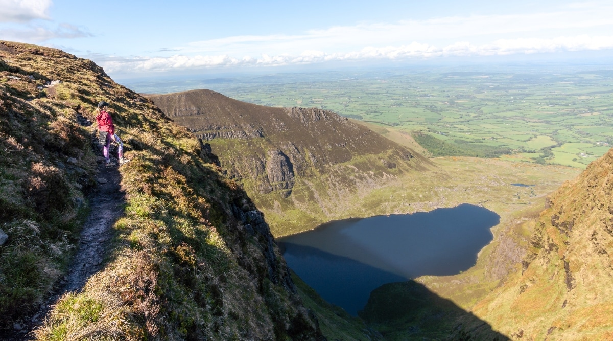 A photograph showcasing Coumshingaun Lake in County Waterford. The image captures the lake's tranquil waters surrounded by hilly terrain. The landscape is characterized by natural greenery and the calm expanse of the lake.