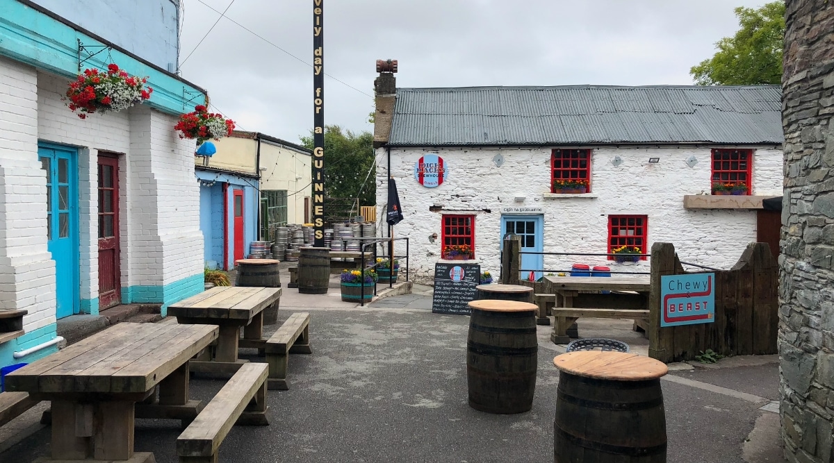 A view capturing a small two-storey building identified as Dick Mack's in Dingle, County Kerry. The exterior features a sign indicating the establishment's name. In the foreground, wooden benches, tables, and stands resembling barrels are arranged, suggesting outdoor seating.