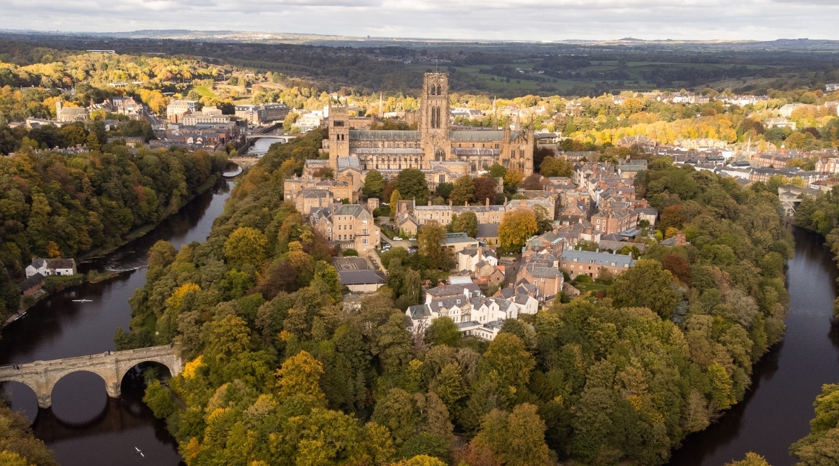 An aerial view capturing Durham City, where Durham Cathedral is prominently featured from the air. The cathedral is surrounded by trees displaying autumnal hues. The overall scene offers a seasonal perspective on the city's architecture and natural surroundings.