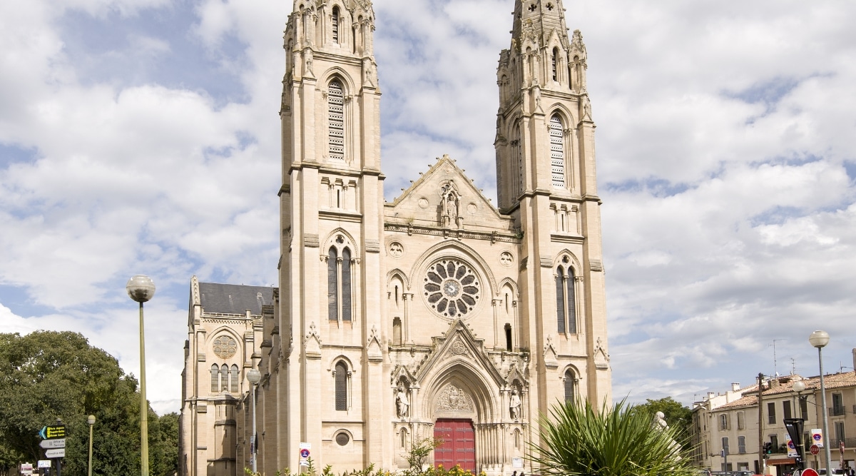 A photograph depicting Eglise Sainte-Baudile in Nimes. The image captures the exterior of the church, showcasing its architectural elements and subdued color palette. The surroundings suggest a typical urban setting with minimal activity around the church