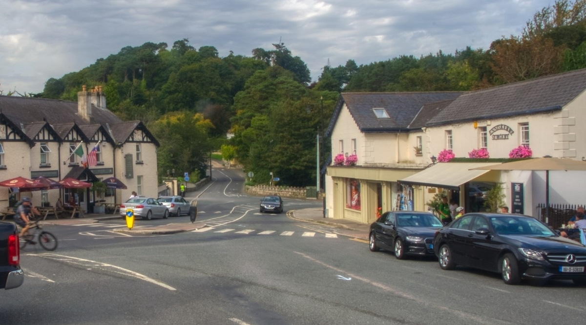 A photograph depicting a street in Feakle, County Clare, Ireland. The image features several buildings along the street, with parked cars lining the roadside. In the background, dense green trees provide a natural backdrop, while the overcast sky contributes to the subdued atmospheric conditions.