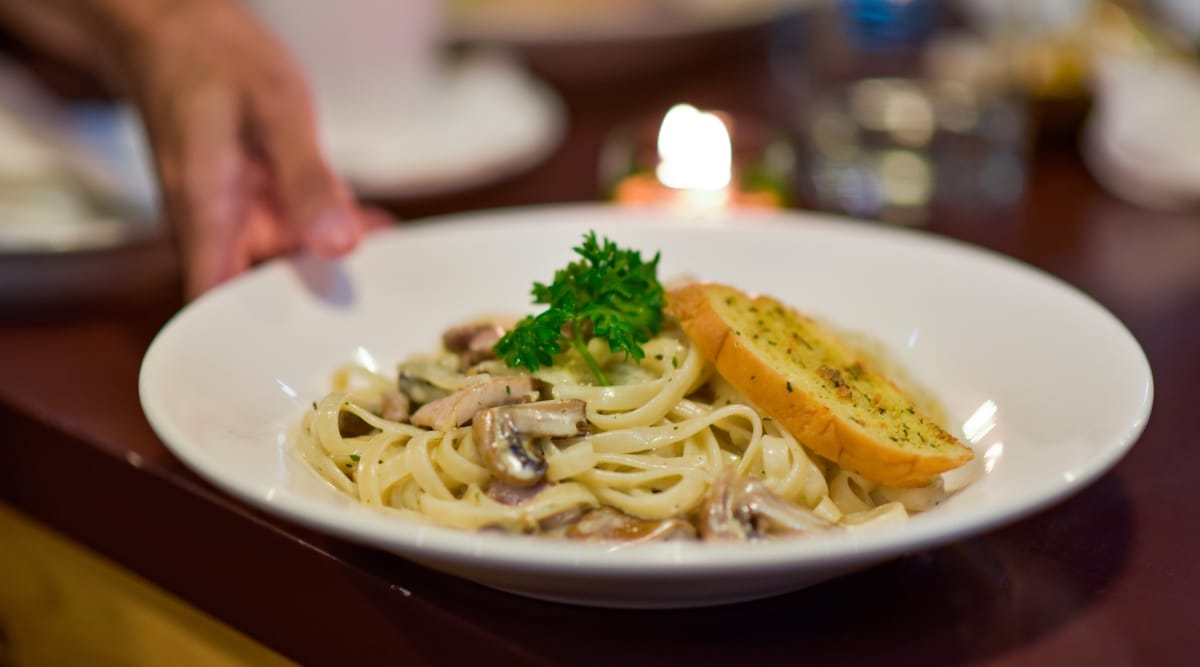 A waiter serves a dish Fettucini carbonara with mushrooms in a white plate in a restaurant. This dish presents as a luxurious and comforting dish, featuring thick ribbons of al dente pasta coated in a creamy, golden-yellow sauce. The slices of tender mushrooms nestled among the pasta.