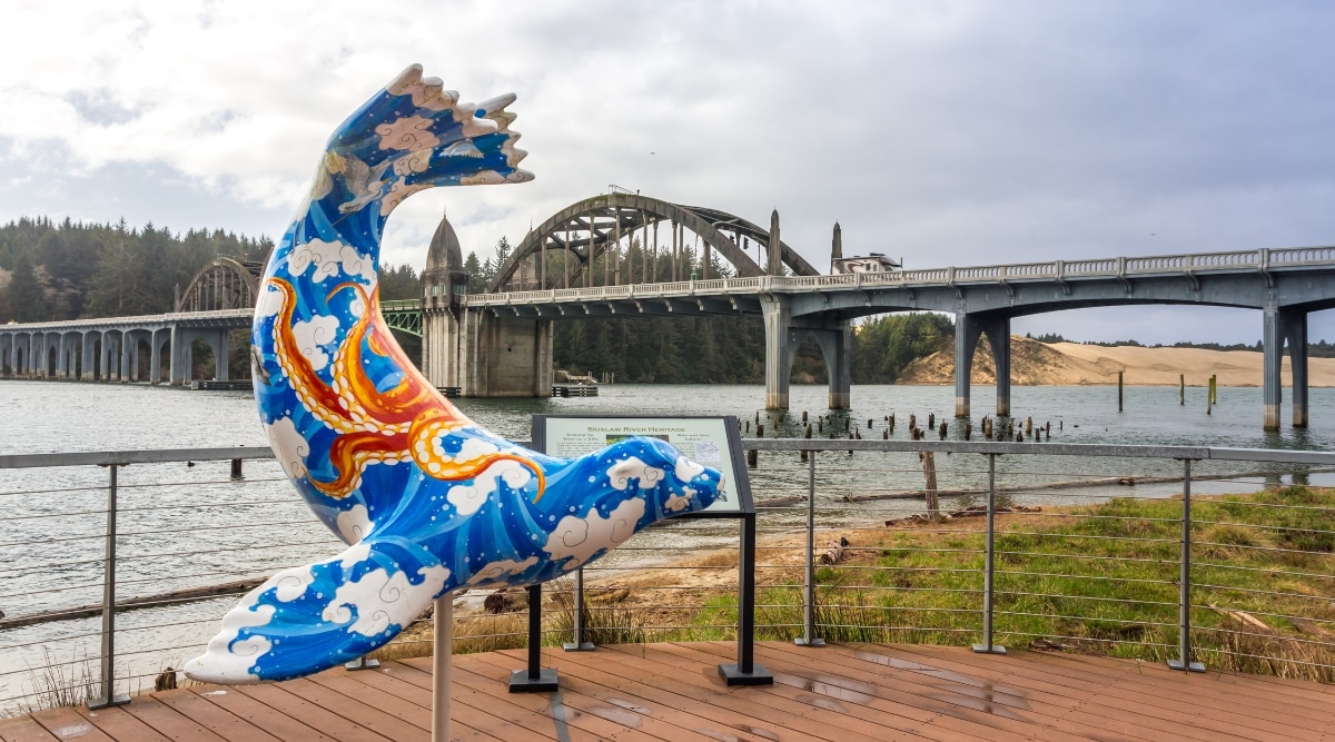 A view of the bridge in Florence, Oregon, featuring a predominantly blue statue of a seal with an orange osmenogus pattern in the foreground. The overcast sky with clouds provides the atmospheric backdrop to the scene. The bridge spans across the waterway.