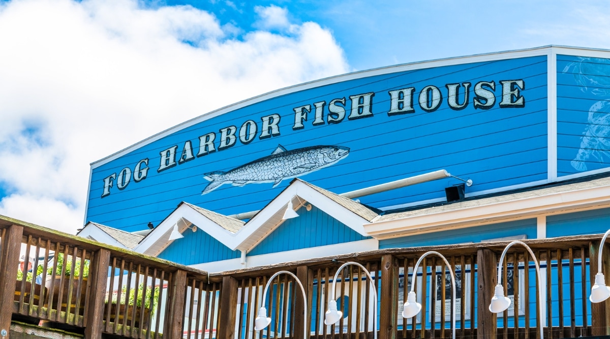 The side of the Fog Harbor Fish House restaurant painted bright blue with a fish underneath and a wooden deck in front of it.