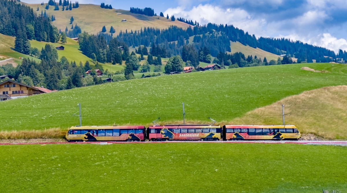 A photograph capturing a Golden Pass color train traversing the Swiss Alps landscape in Gstaad, Switzerland. The image showcases the train's journey against the mountainous backdrop, highlighting the scenic beauty of the region.