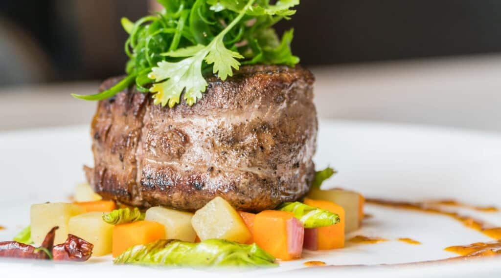Close-up of beef steak with stewed vegetables on a white plate in a restaurant. The piece of steak is thick, juicy, brownish in color with a slightly crispy crust. The steak is topped with fresh cilantro sprigs. Vegetables include carrots, zucchini, onions and lettuce.