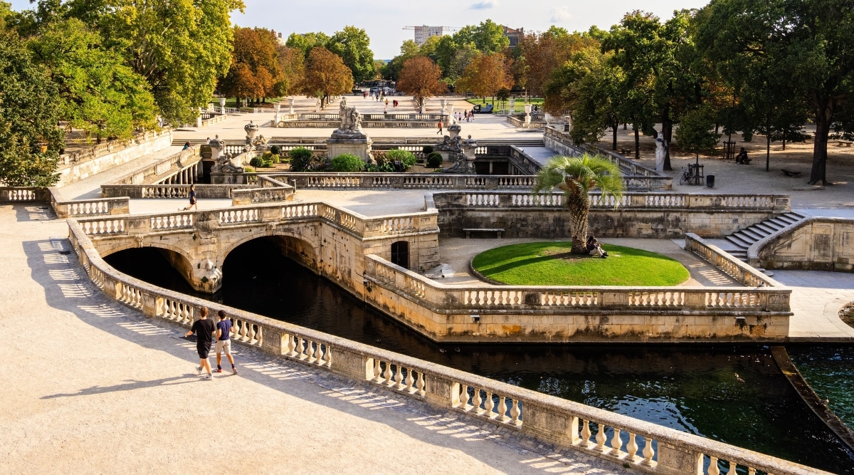 A photograph capturing Jardins de la Fontaine in Nimes. The image showcases the formal gardens featuring pathways, manicured lawns, and classical sculptures.