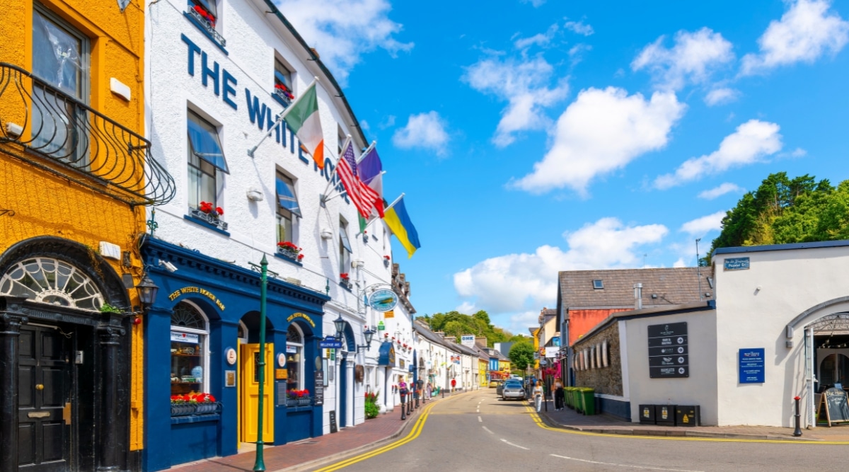 A photograph capturing Market Street in Kinsale, County Cork, Ireland. The image showcases a row of colorful shops lining the main shopping street of this historic port and fishing town. The architectural elements of the buildings reflect a mix of traditional and contemporary styles.