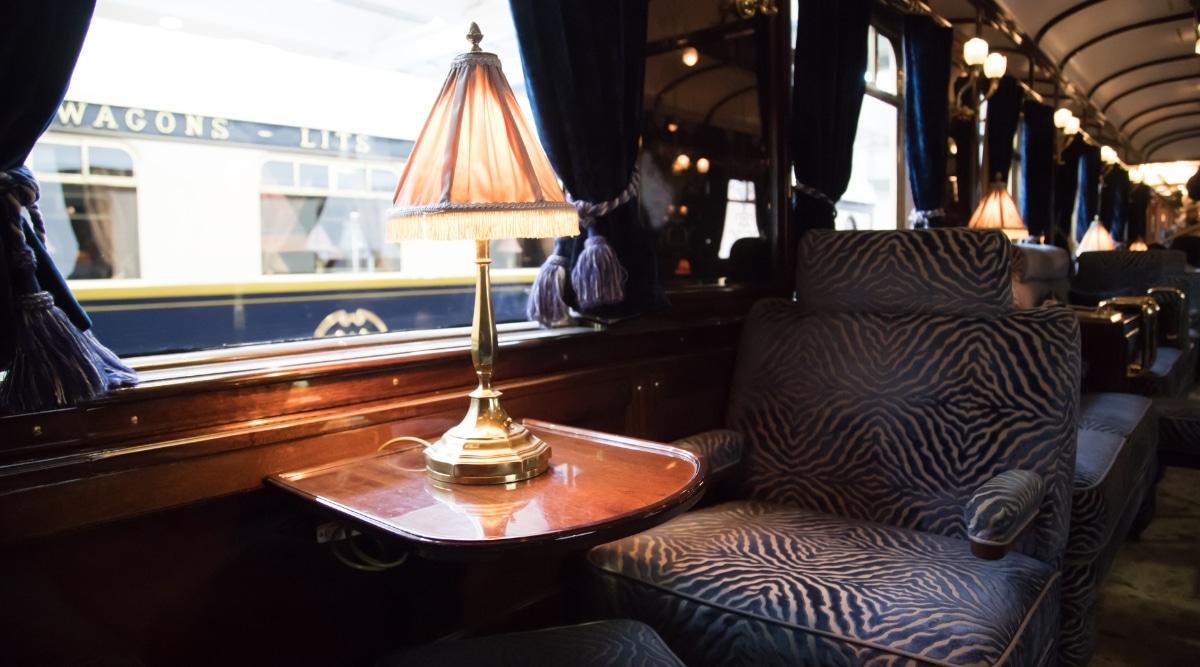 A close-up view of the interior of a train compartment featuring a floor lamp standing on a wooden table. The image captures a large soft chair, adorned with blue curtains, and offers a glimpse of another train through the window. The scene is devoid of people, maintaining a sense of tranquility.