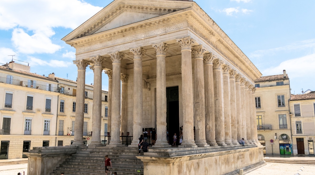 A photograph capturing La Maison Carrée in Nimes, France. The image showcases the well-preserved Roman temple, known for its classical architecture. The façade features prominent columns and intricate details characteristic of ancient Roman design. The surrounding area appears relatively quiet, with a few visitors observing the historical monument.
