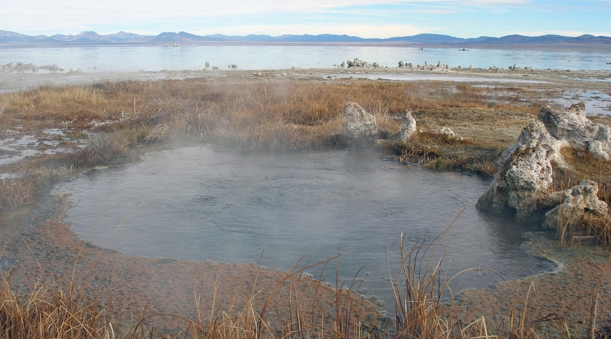 A photograph depicting Mono Lake Hot Springs in California. The image captures the natural geothermal features of the hot springs, situated in proximity to Mono Lake. The landscape surrounding the hot springs is characterized by arid terrain and sparse vegetation. 