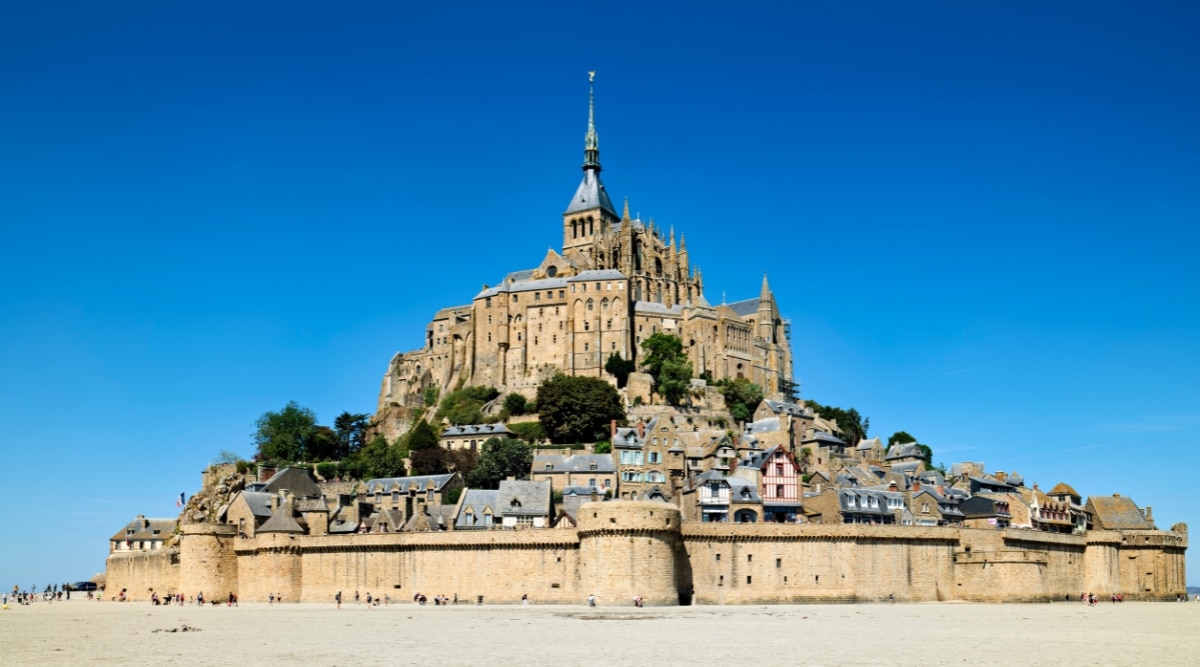 A direct view of Mont Saint Michel on a clear and bright day, featuring a cloudless sky. Numerous people are visible in the vicinity. The image captures the architectural prominence of Mont Saint Michel, a renowned historical abbey and tourist attraction.