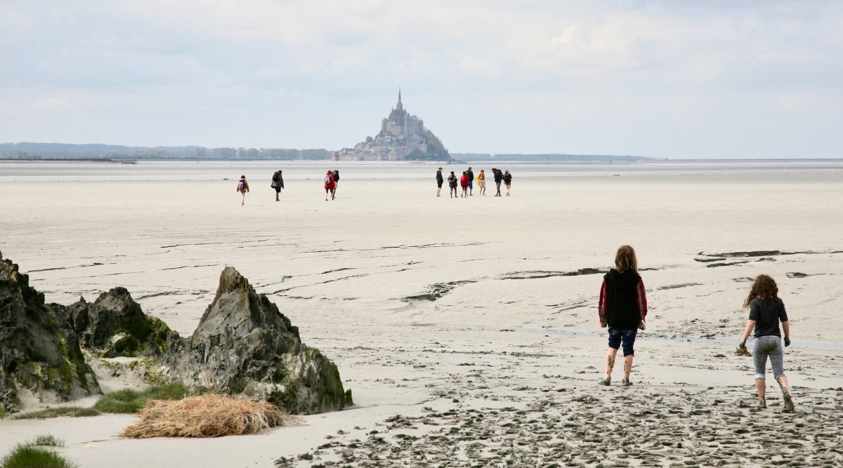 Mont Saint Michel low tide on a cloudy day as tourists walk through the muddy sandy silt.