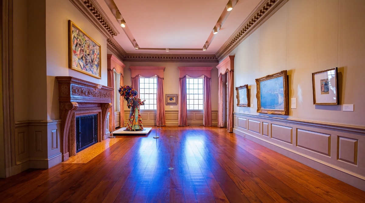 An image capturing a museum hall with beige walls and a wooden floor. Paintings are displayed on the walls throughout the empty hall, devoid of any visitors. 