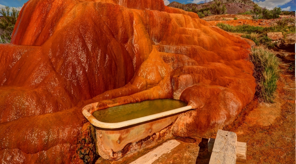 A view of Mystic Hot Springs in Utah, showcasing a red stone formation surrounding a bathtub filled with water. The geological features of the red stone are prominent, creating a visually distinctive setting for the hot springs. The bathtub is positioned within the natural surroundings, contributing to the overall aesthetic of the location.