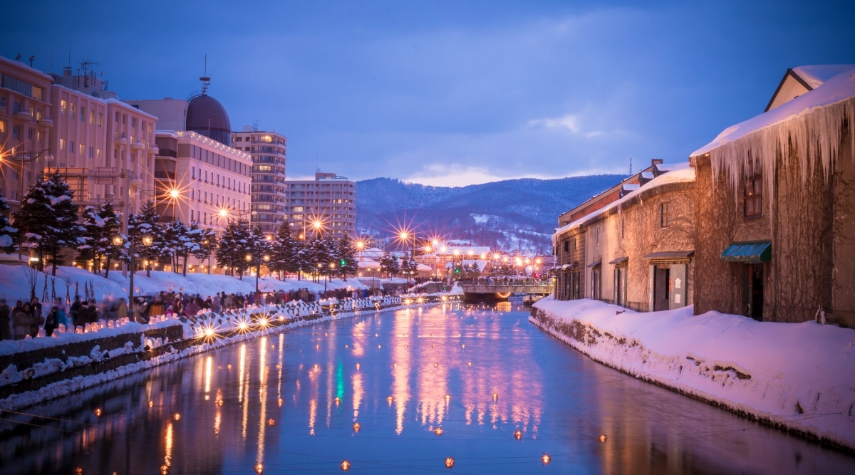 A photograph capturing the winter scene of Otaru's 'Way of Snowlight.' The image depicts a snowy landscape featuring a river flanked by houses on both sides. Numerous lights are visible, illuminating the surroundings. 