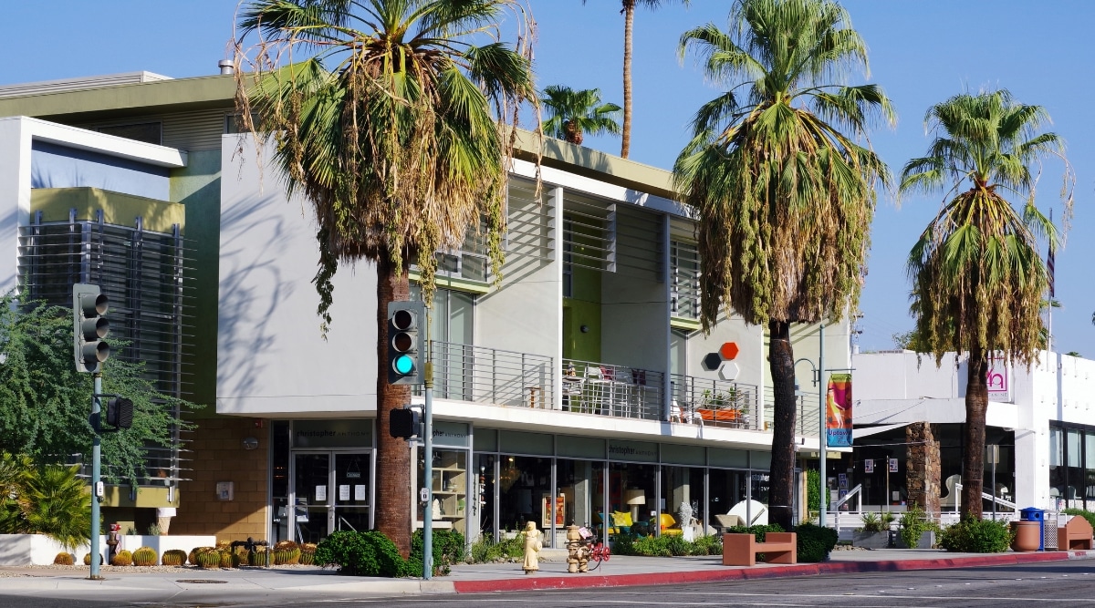 A photograph depicting North Palm Canyon Drive in Palm Springs, California, featuring a mix of commercial and residential buildings. The image showcases the urban landscape with a row of structures along the street.