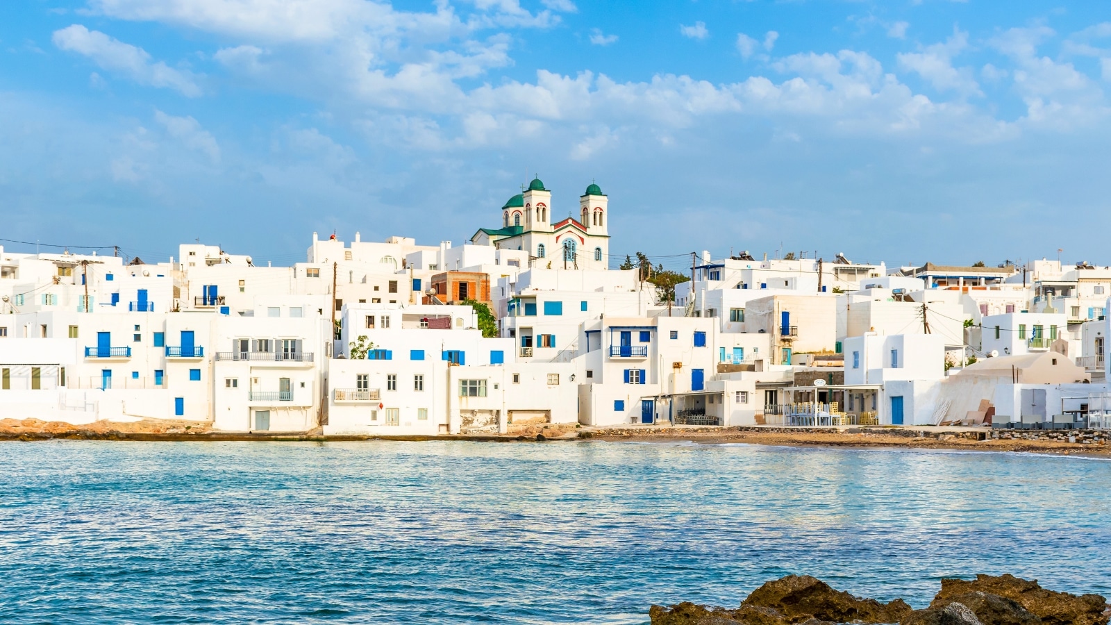 A scene depicting the traditional Cycladic architecture in Naousa, Paros. The image captures whitewashed houses, a hallmark of the region, with the Christian church of Kimisis Theotokou as a prominent feature. The structures are arranged in the characteristic style of Cycladic villages, creating a visually cohesive landscape.