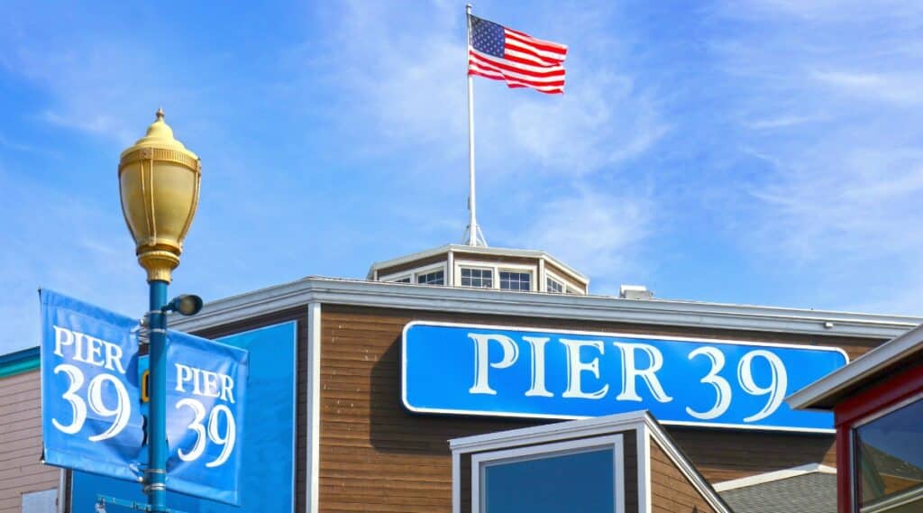 Sunny day shot of the Pier 39 sign with an American Flag raised and waving above it and a lamp post in the left foreground also with banners advertising Pier 39.
