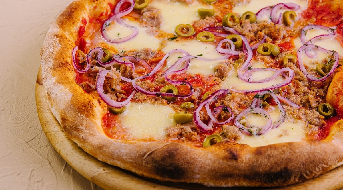 Close-up of Pizza with tuna and red onion on a serving board. The surface is adorned with generous flakes of canned tuna, red onion slices, green olives and lots of melted cheese. The pizza crust is slightly crispy and golden brown.