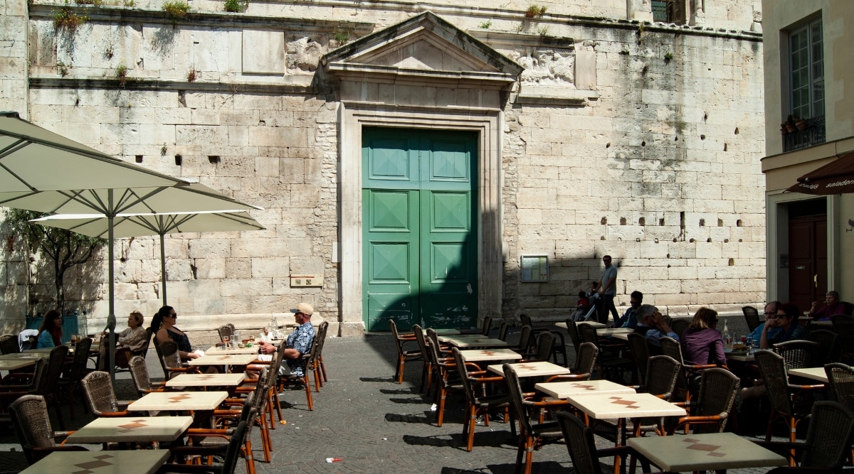 A photograph capturing Place aux Herbes in Nimes, France. The image depicts the square's architectural layout, with buildings lining its perimeter.