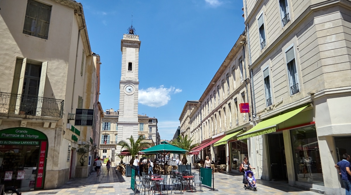 A photograph depicting Place de l'Horloge in Nimes. The image showcases the square's architectural elements, featuring surrounding buildings, facades, and the central clock. Numerous individuals can be observed in the square.