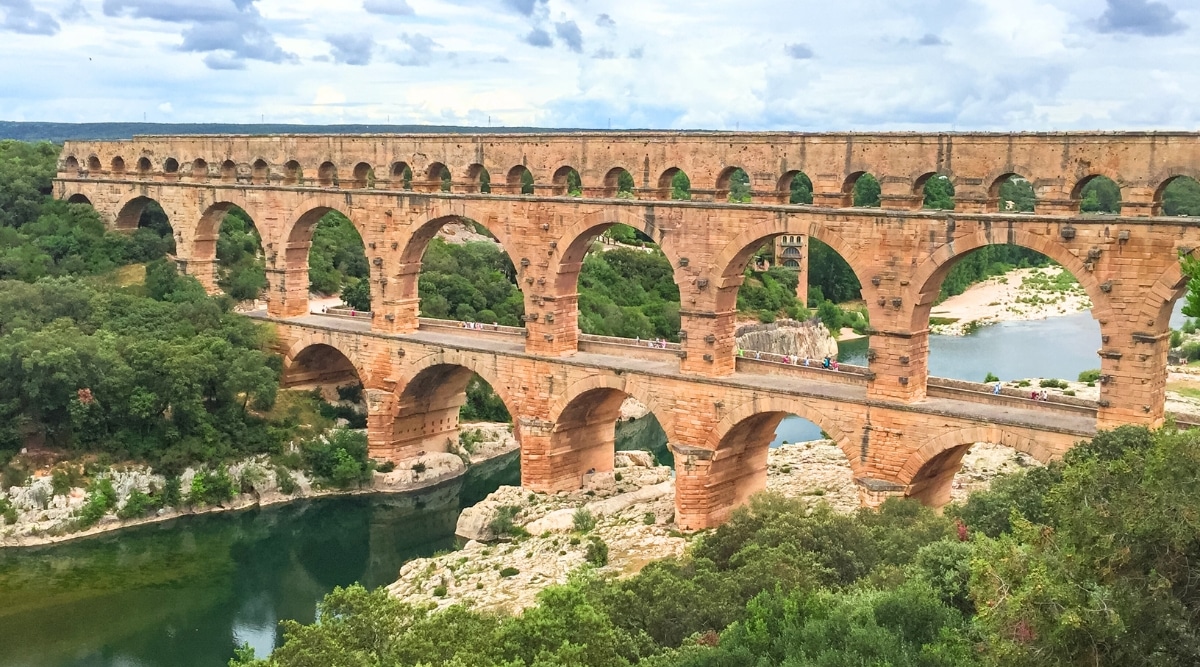 A photograph capturing Pont du Gard, a historical Roman aqueduct bridge in Nimes, France. The image showcases the well-preserved three-tiered arches spanning the Gardon River. The surrounding landscape features natural elements and the clear, blue sky.