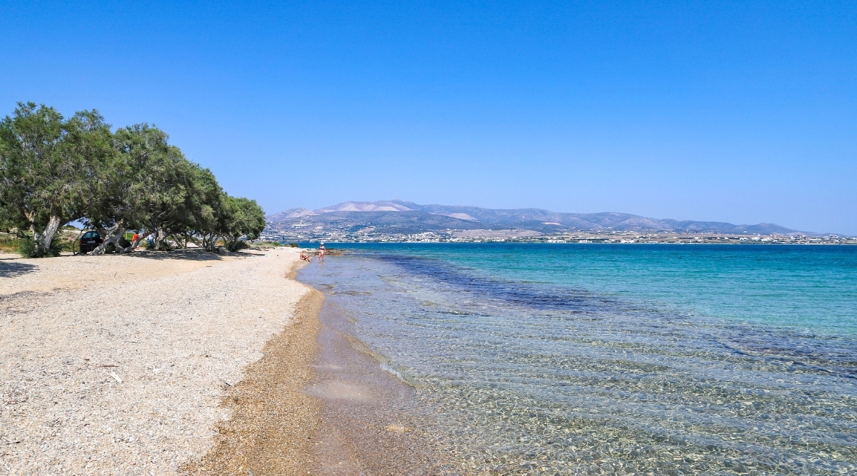 A photograph capturing Psaralyki 1 Beach in Antiparos, Cyclades, Greece. The image showcases crystal-clear waters and golden sandy shores, characteristic of the beach's natural features. Shadowy trees line the coastline, providing areas of shade for visitors
