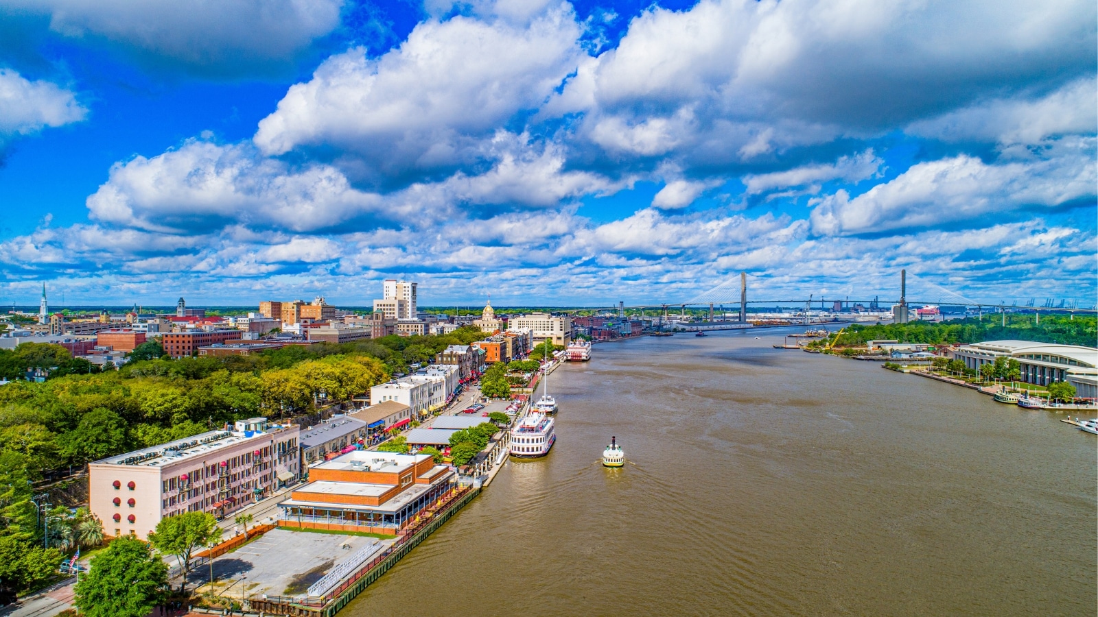 An aerial photograph capturing the downtown skyline of Savannah, Georgia, along the Savannah River. The image provides a comprehensive view of the city's architectural landscape, featuring a mix of buildings and structures.