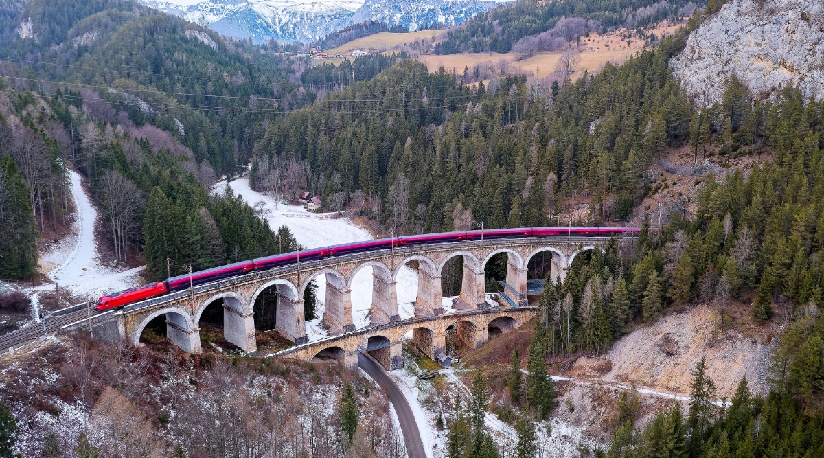 The image depicts an expansive railway bridge, a part of the UNESCO World Heritage Site Semmering Railway in Austria. A lengthy red train, comprising numerous carriages, travels along the tracks spanning the entire length of the bridge. The surroundings feature a vast territory with mountains, hills, and forests, emphasizing the scenic landscape surrounding the railway infrastructure.