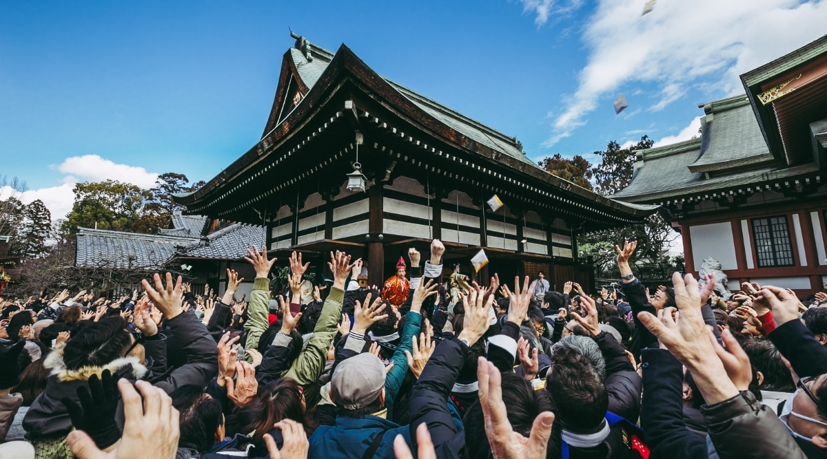 A crowded scene capturing a Setsubun event, where numerous individuals are raising their hands towards the top on a bright day.