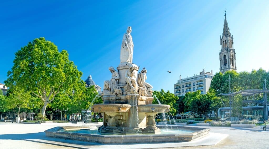 A close-up photograph featuring the Fontaine Pradier in Nimes. The image showcases the architectural details of the fountain, located within Square de l'Esplanade. The fountain, known as Fontaine Pradier, is depicted against a neutral urban backdrop.