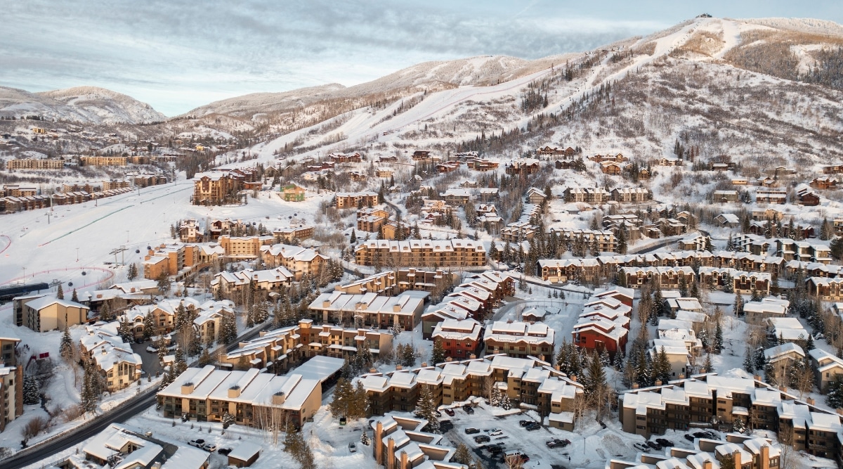 A drone view capturing the snowy cityscape of Steamboat Springs. The image depicts an extensive residential area with snow-covered rooftops, showcasing the winter conditions prevalent in the region. Numerous houses are visible throughout the landscape, surrounded by a blanket of snow. The background features snow-covered hills.