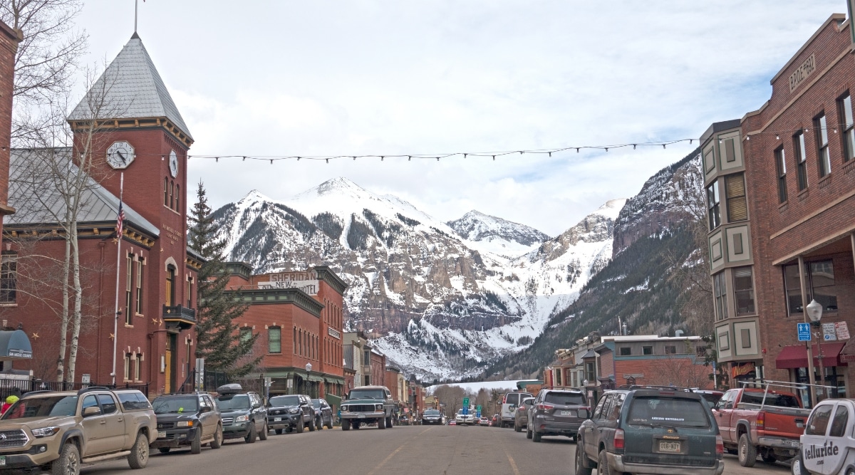  A ground-level photograph capturing a street view in Telluride, Colorado. The image features a building flanked by parked cars on both sides of the street. The overcast sky and the absence of leaves on the trees indicate the winter season. Directly across the street, a clear view of snow-capped mountains is prominent in the background.