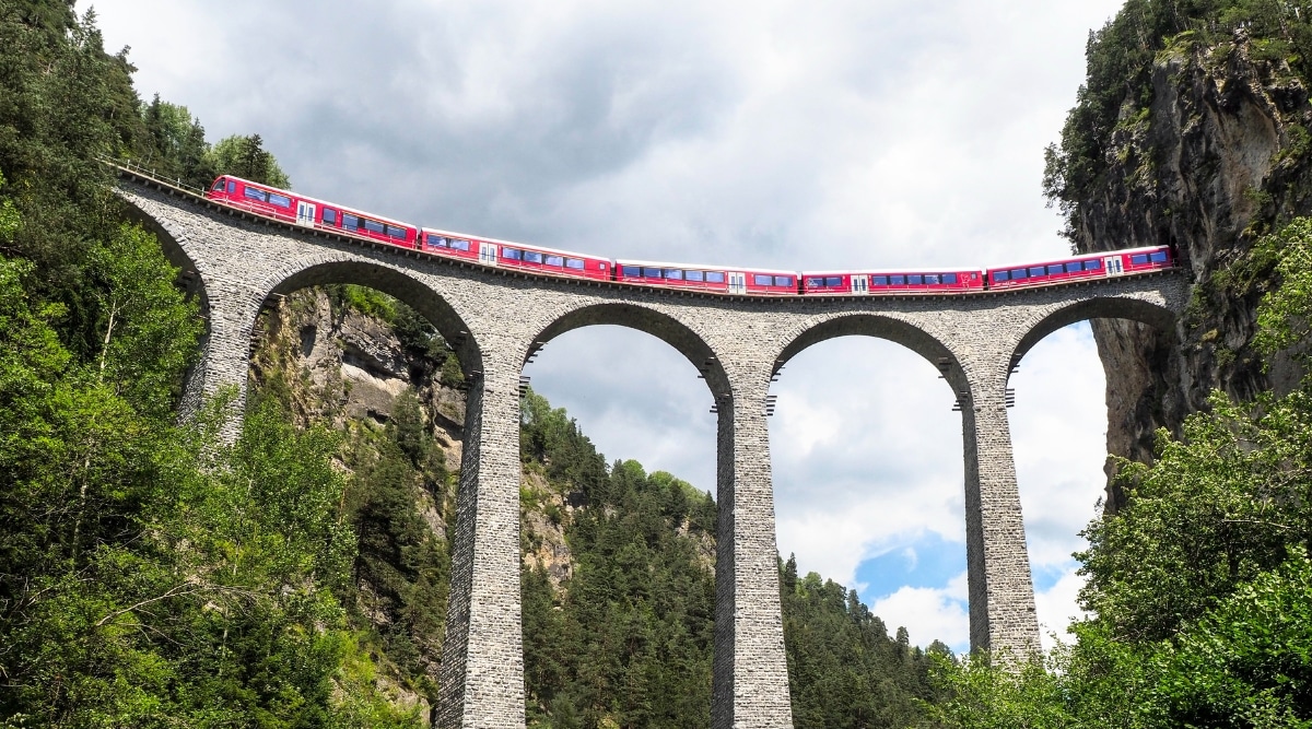 A photograph capturing the Viaduct of the Rhaetian Railway in Filisur, Switzerland. The image showcases the red and white train in motion, traversing the historic railway viaduct.