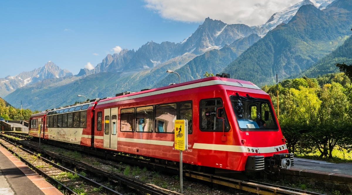 The image captures a red train consisting of 2 carriages in motion along the Mont Blanc Express in France. The backdrop features picturesque mountains, contributing to the scenic landscape.