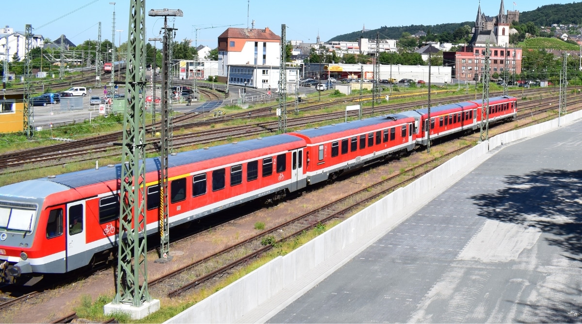 The image captures a red train consisting of four carriages in motion along the tracks of the Rhine Valley Railway in Germany. In the background, a town is visible.