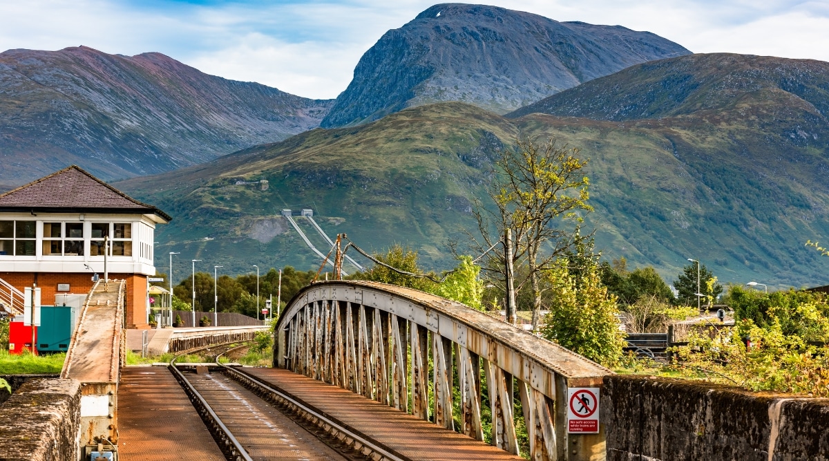 The image features a straightforward representation of the railway infrastructure, with the tracks extending across the bridge. The backdrop showcases the Ben Nevis mountains, providing a scenic context to the railway setting.