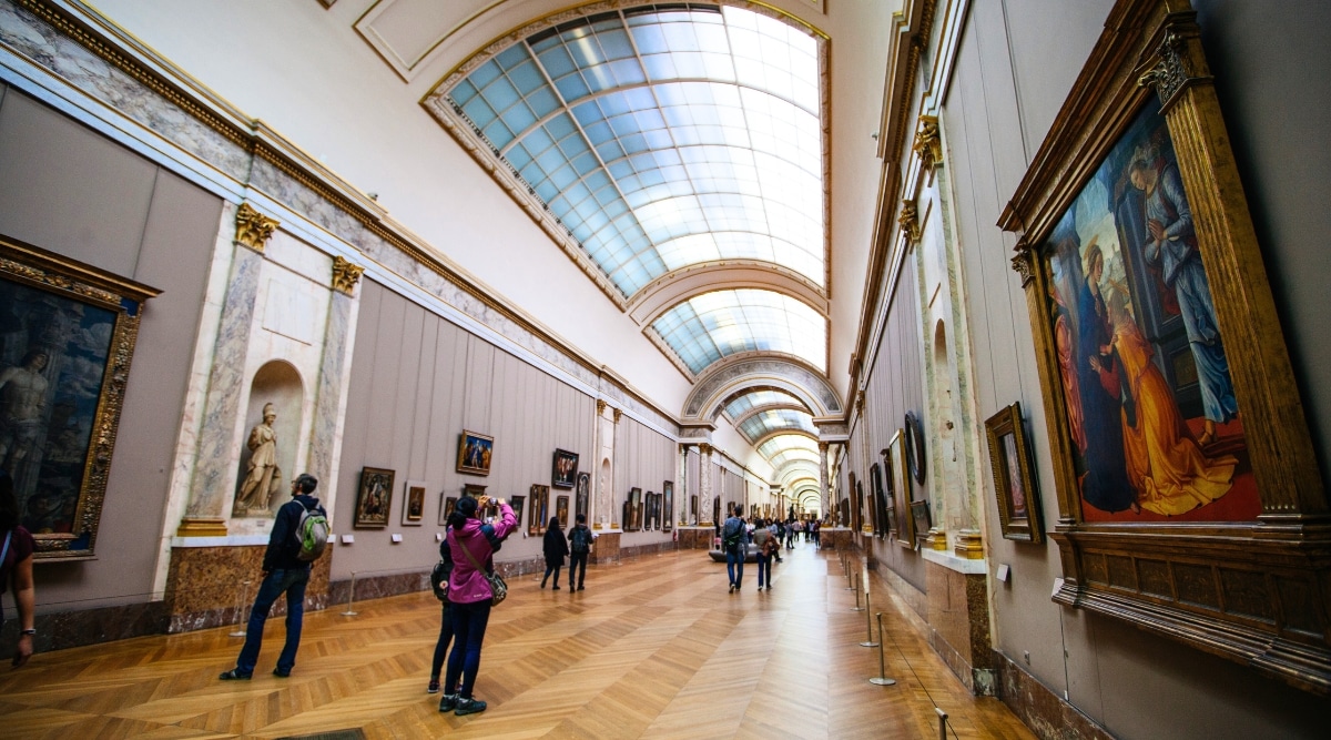 A photograph capturing visitors walking through a long, well-lit room within the Louvre museum. The room features paintings on the walls and a glass ceiling, contributing to the ample illumination of the space.