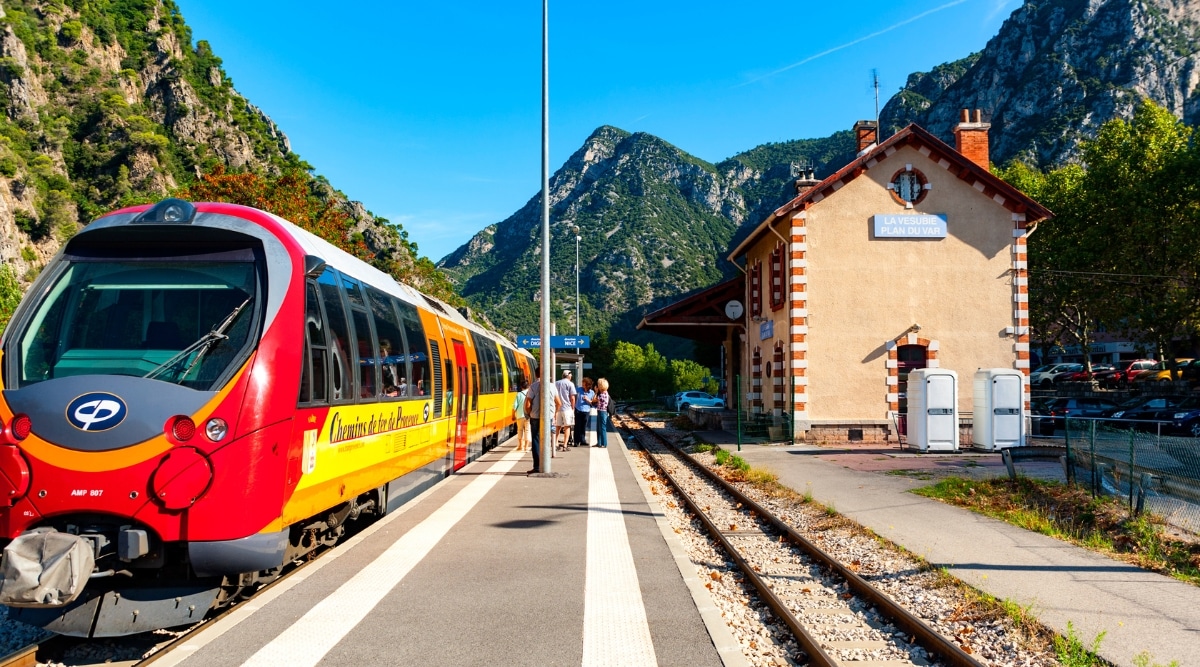The image depicts a modern red-yellow track train at the Train des Pignes station in France. Passengers are seen boarding the train. Adjacent to the train stands a two-storey station building.