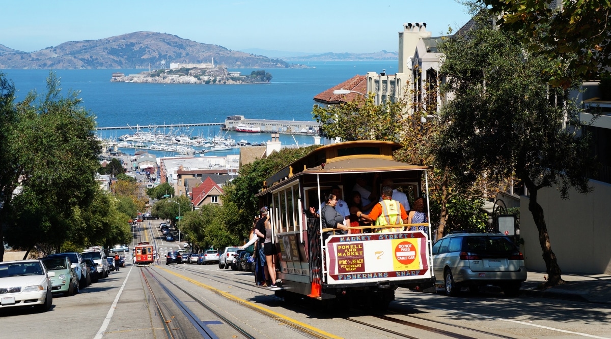Tourists riding one of San Francisco's famous trolley cars down hill towards the wharf with Alcatraz Island visible in the background on a clear sunny day.
