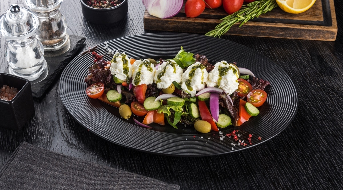A photograph showcasing a vegetable salad presentation on a black tray, featuring a topping of feta mousse. The image highlights the culinary composition with a focus on color contrast and presentation. The assortment of vegetables appears fresh and neatly arranged. The feta mousse adds a creamy texture, contributing to the overall aesthetic appeal of the dish.