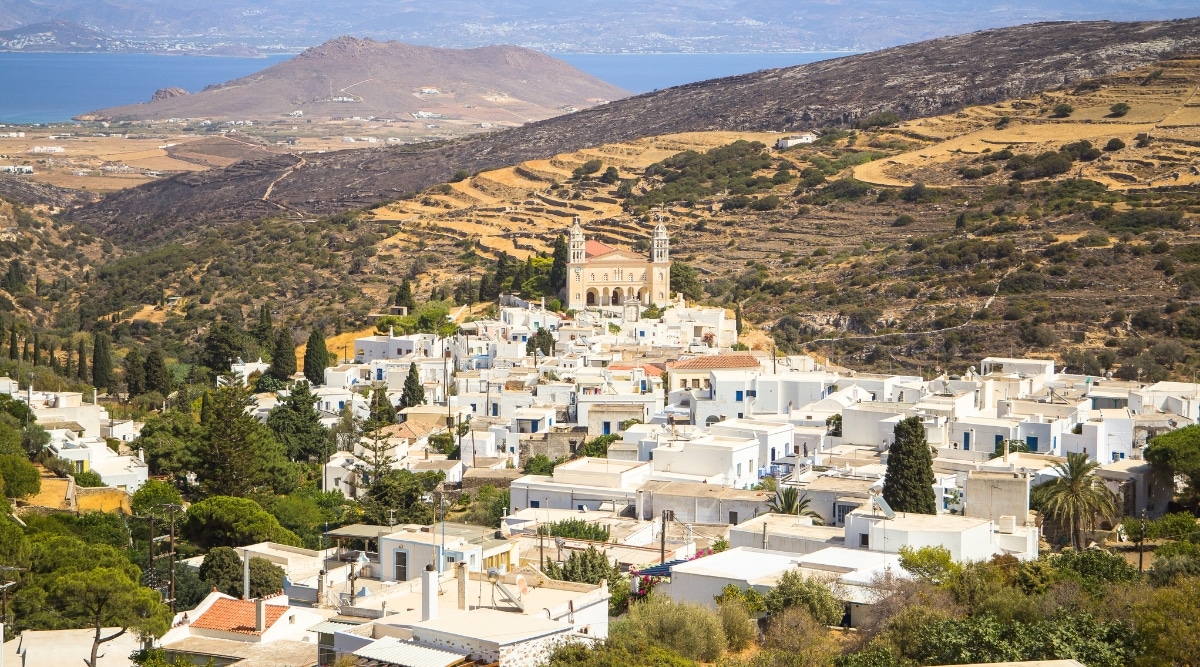 An aerial view overlooking the traditional village of Lefkes and Agia Triada Church in Paros, Greece. The image captures the characteristic architecture of the village, with traditional white-washed buildings and the church's distinct features. In the distance, the island of Naxos is visible, contributing to the scenic backdrop.