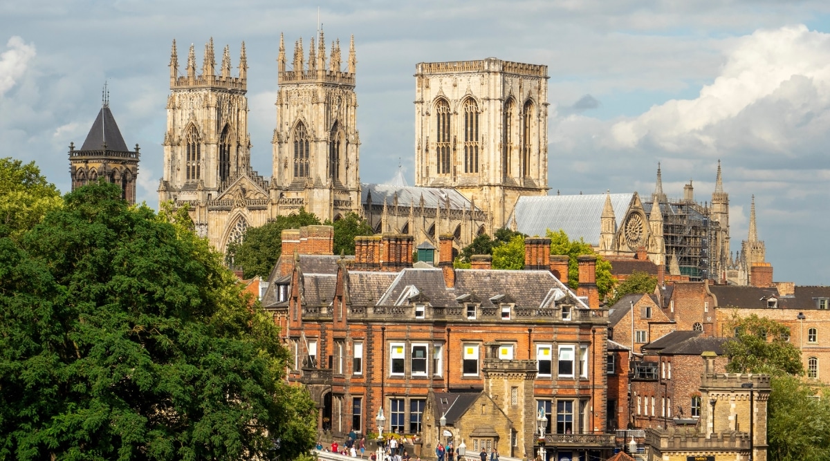 A photograph capturing York Minster as seen from the city walls over rooftops in York, Yorkshire. The image showcases the iconic cathedral bathed in sunshine, with surrounding rooftops forming the foreground. The architectural details of York Minster are visible in the sunlight, highlighting its historical significance.
