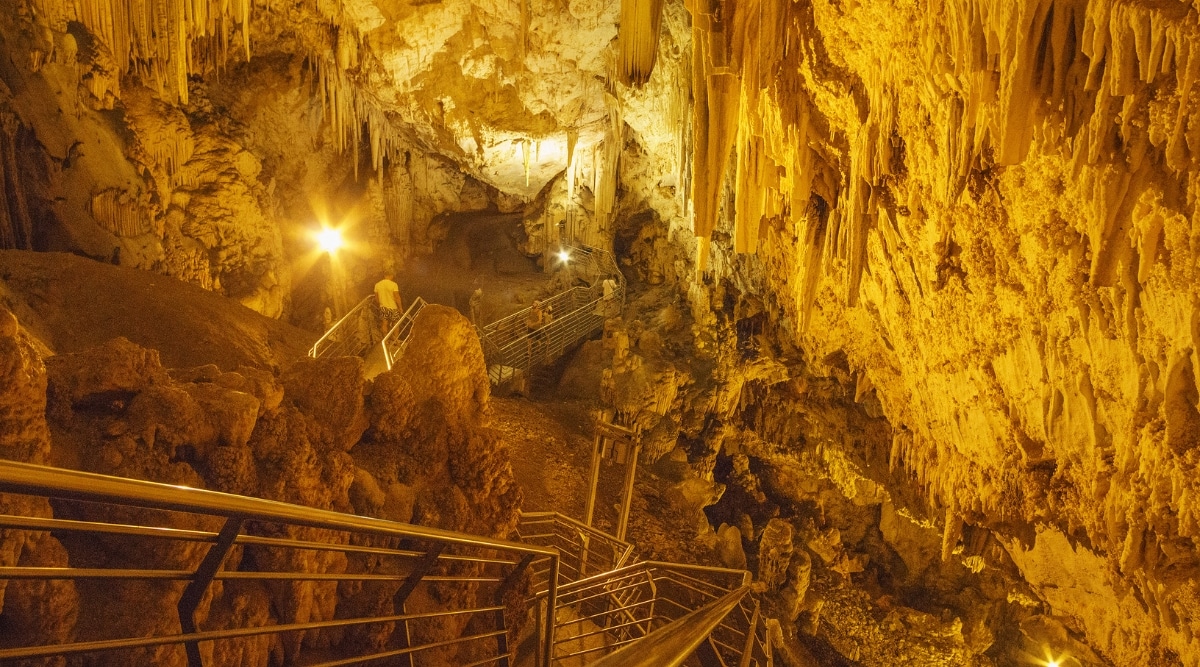 A photograph capturing the famous cave of Antiparos in the Cyclades, Greece. The image showcases the expansive and atmospheric cave entrance, featuring a descent of 300-plus concrete steps into the well-lit interior.