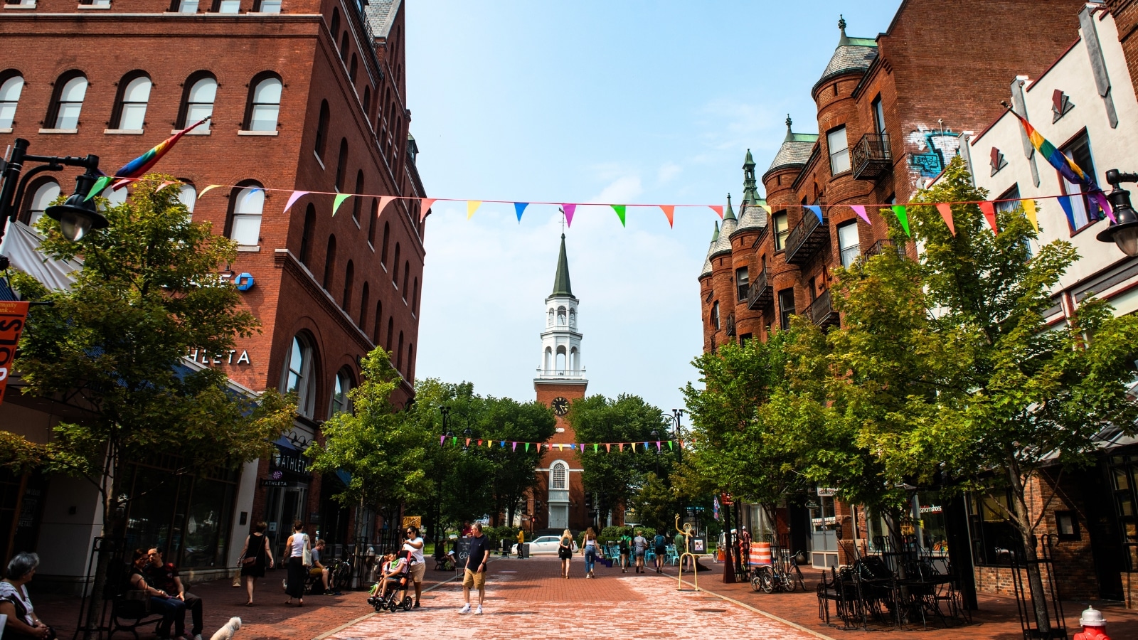 A photograph depicting The Church Street Marketplace in Burlington, Vermont. The image showcases the pedestrian-friendly street with shops and commercial establishments lining both sides. People are visible engaging in typical weekday activities, such as shopping or strolling along the brick-paved walkway.
