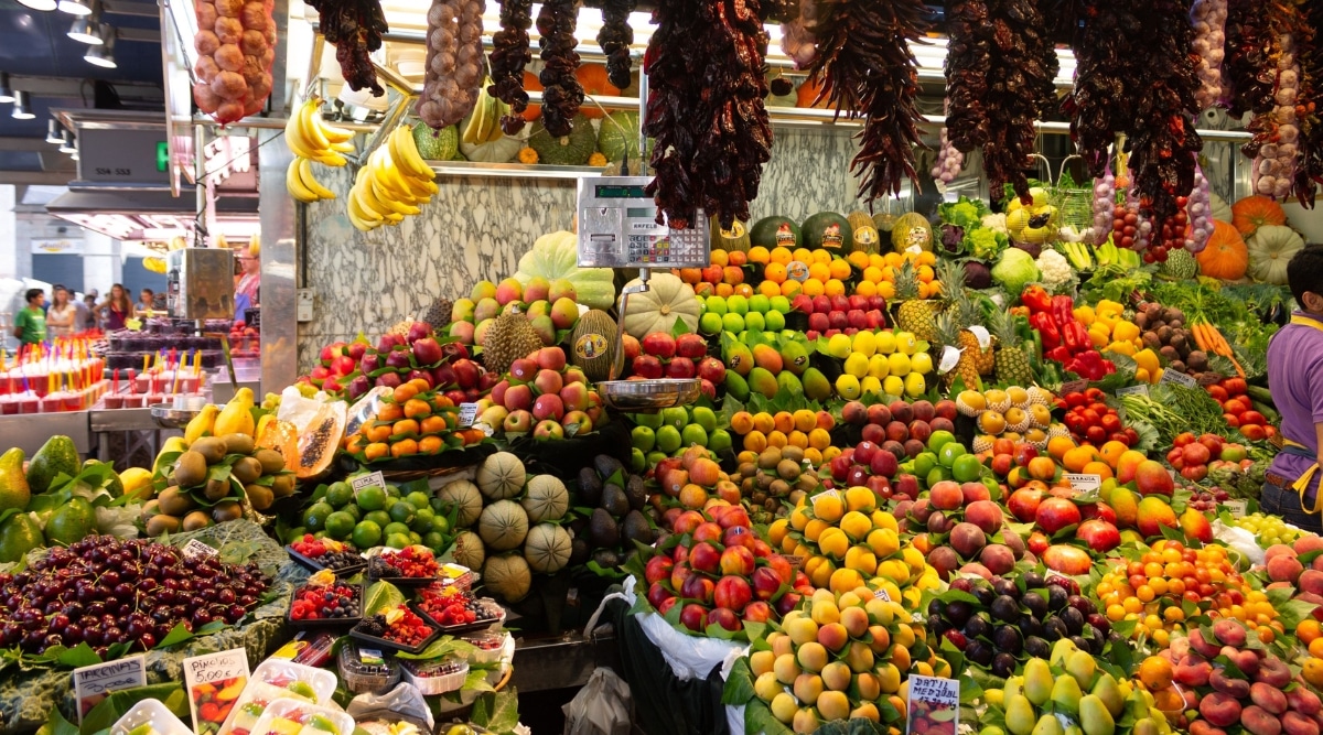 A photograph depicting a fruit stand within a covered market. The image showcases a variety of fruits neatly arranged and displayed for sale. The stand appears to be part of a larger market setting, emphasizing the availability of fresh produce.