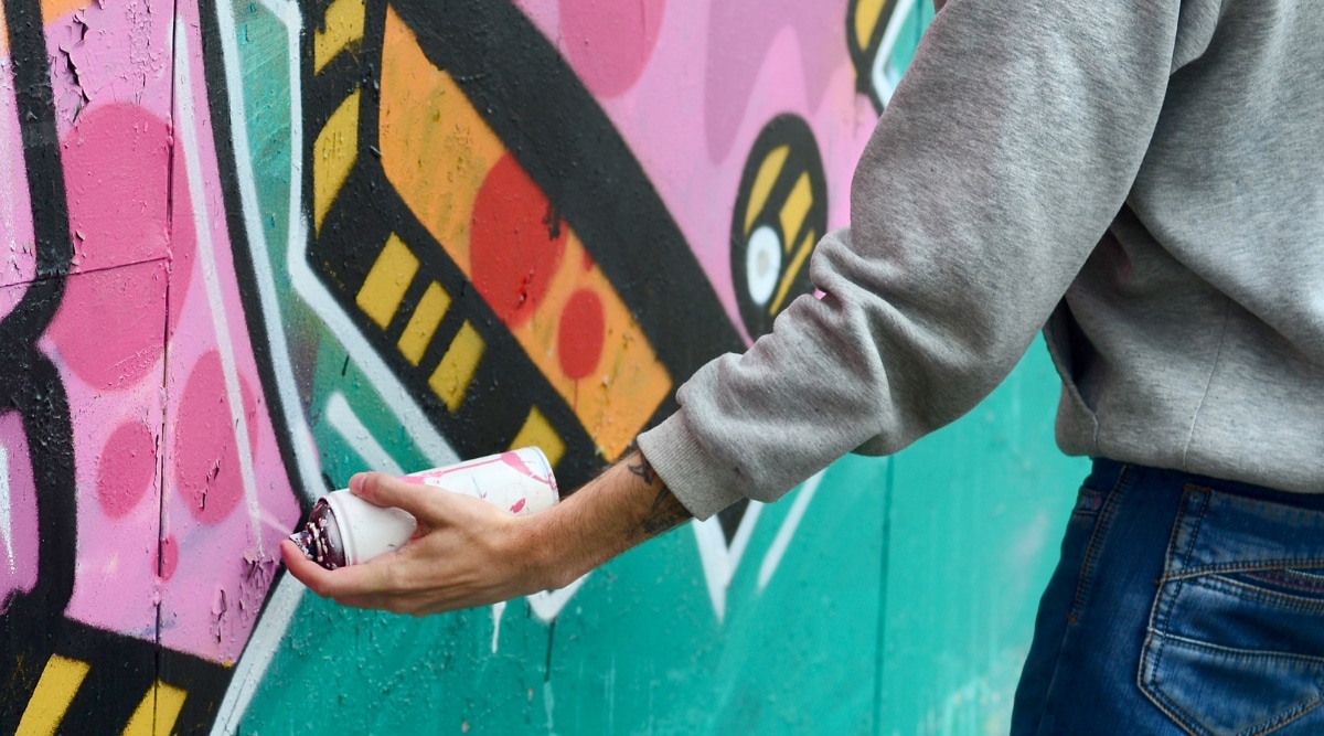 A photograph depicting an individual painting graffiti on a wall using a spray can. The image captures the artist engaged in the act of creating street art.