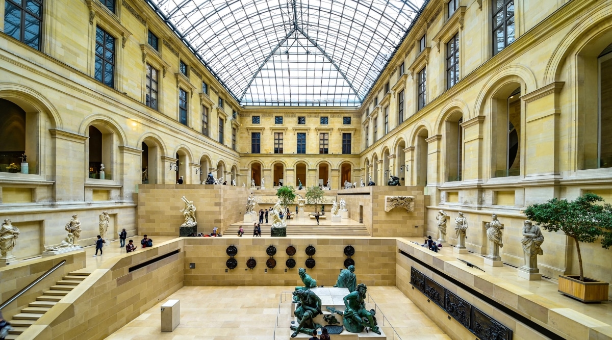 An inner view of the Louvre Museum hall in Paris, featuring sculptures and visitors. The image showcases the interior space filled with various sculptures, and visitors can be observed exploring the exhibits.